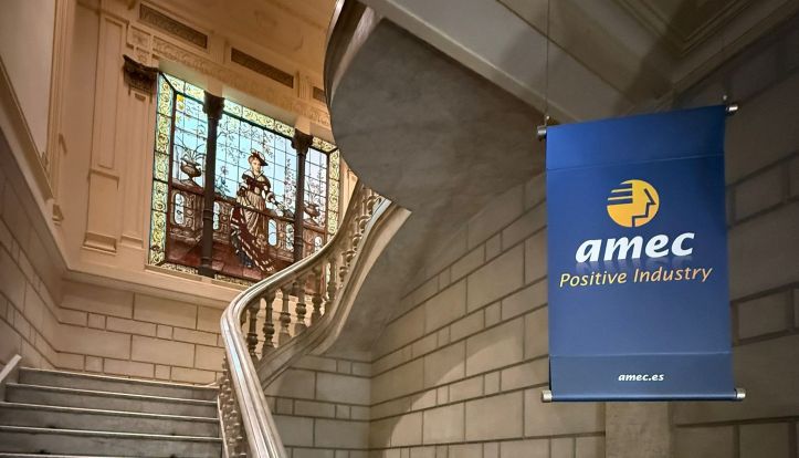 Acceso a las instalaciones de AMEC en la Gran Vía de las Cortes Catalanas en Barcelona Logotipo de la iniciativa Positive Industry de AMEC ubicado en un pasillo arquitectónico con un ventanal de vitral al fondo.